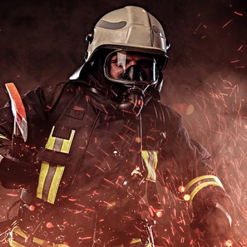 A firefighter dressed in a uniform and an oxygen mask holds a red axe standing in fire sparks and smoke over a dark background.